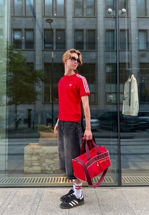 Camiseta roja de manga corta con rayas blancas, pantalones cortos de mezclilla negros hasta la rodilla y una bolsa de deporte roja. Zapatillas negras y calcetines blancos con rayas rojas.