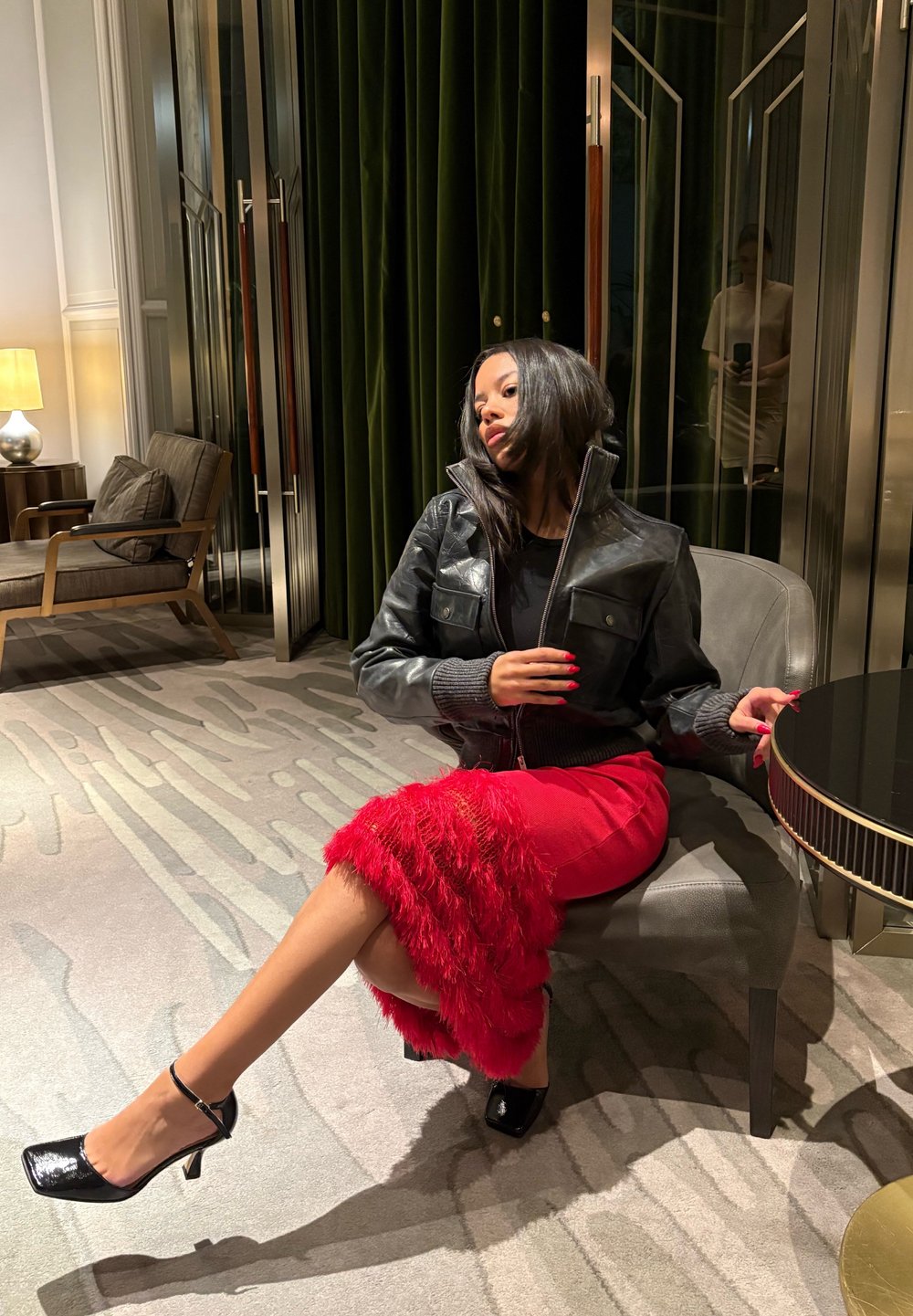 Casaco de couro preto com detalhes em malha cinza, saia vermelha com acabamento em penas e saltos pretos de verniz. Cenário em interior moderno com iluminação suave.
