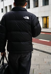 Μαύρο μπουφάν πούφ με ψηλό γιακά και ορατές ραφές. Λογότυπο "THE NORTH FACE" σε λευκό στην επάνω πλάτη. Σκούρο γκρι τζιν και τσάντα.