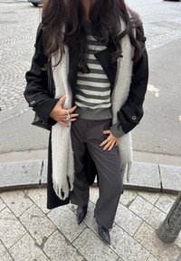 Črni plašč, sivo črtast pulover, sive hlače, dolga svetlo siva šal s resicami in črni koničasti škornji. Roke okrašene z obročki.