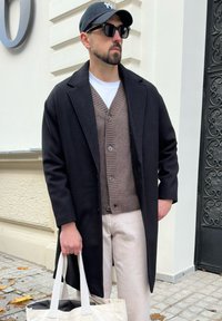 Mann in schwarzem Mantel, braunem Cardigan, weißen Hosen, schwarzer Mütze und Sonnenbrille, der eine weiße Tragetasche am Eingang eines Gebäudes hält.