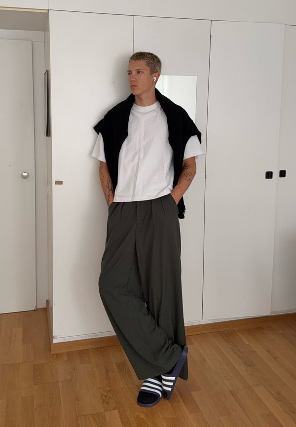 Joven con el pelo corto que lleva una camiseta blanca, un suéter negro colgado sobre los hombros, pantalones oscuros y sueltos y zapatillas de rayas, de pie en el interior.