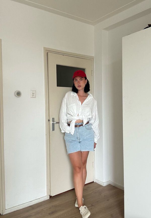 Junge Frau trägt eine rote Kappe, weißes gebundenes Hemd, Jeans-Shorts und weiße Schuhe, steht drinnen in der Nähe einer geschlossenen beigen Tür.