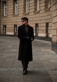 Long manteau noir par-dessus un pull vert, pantalon marron et gants noirs ; entouré de bâtiments en brique avec de grandes fenêtres.
