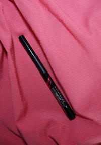 Kajal Maybelline Hyper Precise All Day v elegantnej čiernej tube s ružovými akcentmi, umiestnený na jemnej ružovej látke. Hladký, valcový dizajn.