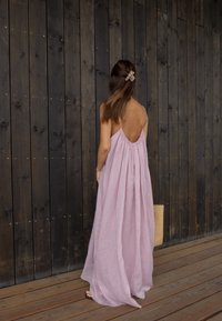 Μάξι φόρεμα από απαλό ροζ λινό, με χαμηλή πλάτη και λεπτές τιράντες. Η ροή της σιλουέτας αναδεικνύεται από μια καφέ τσάντα tote.