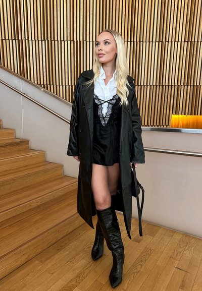 Abrigo de cuero negro, camisa blanca, vestido de encaje negro por encima, botas negras hasta la rodilla con acabado texturizado, de pie en unas escaleras de madera.