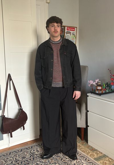 Joven con bigote que lleva una chaqueta de mezclilla negra, suéter a rayas, pantalones anchos negros y zapatos negros, de pie dentro de una habitación cerca de una silla y una cómoda.