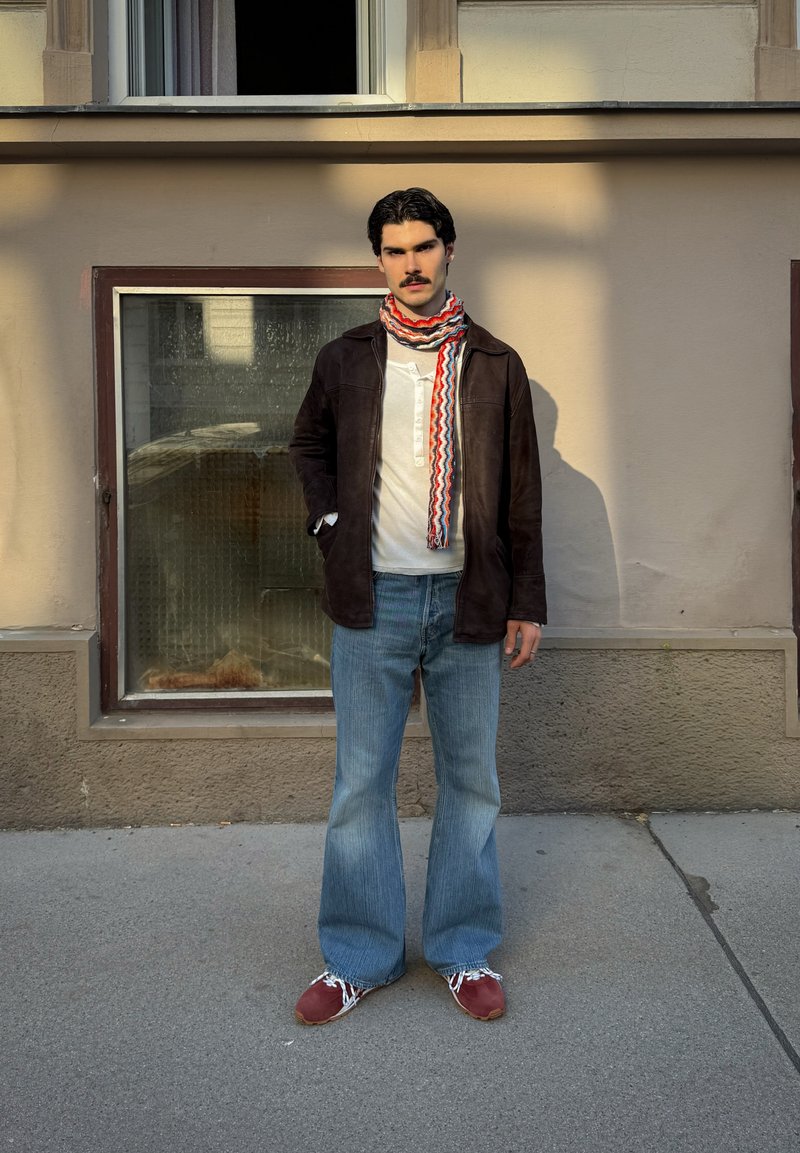 Homme avec moustache portant une veste sombre, une chemise blanche, une écharpe à motifs, un jean évasé et des chaussures rouges, debout sur un trottoir devant un bâtiment.