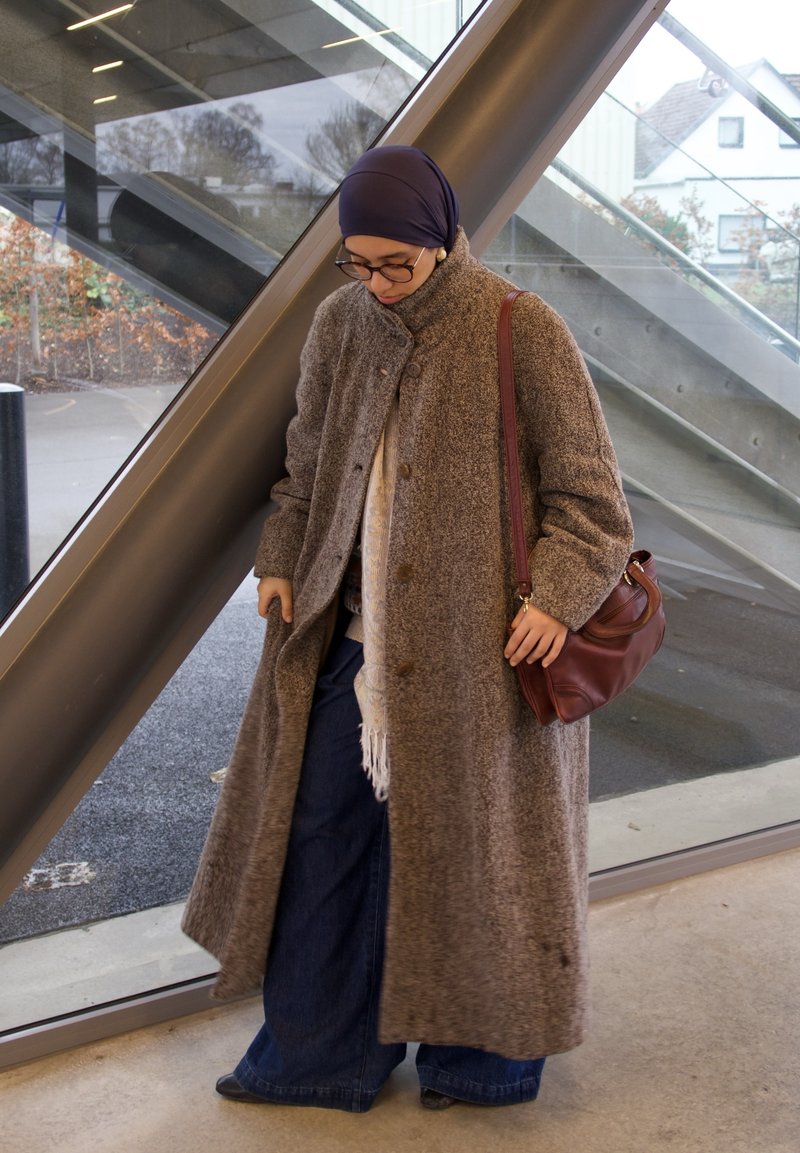 Long manteau brun texturé avec un col montant, porté sur une écharpe à motifs et un pantalon bleu à jambes larges. Tient un petit sac en cuir marron.