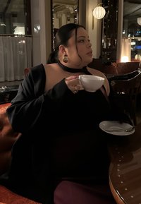 Femme aux cheveux foncés portant un haut noir et des boucles d'oreilles, tenant une tasse à thé blanche avec sa soucoupe, assise dans un café ou restaurant élégant et faiblement éclairé.