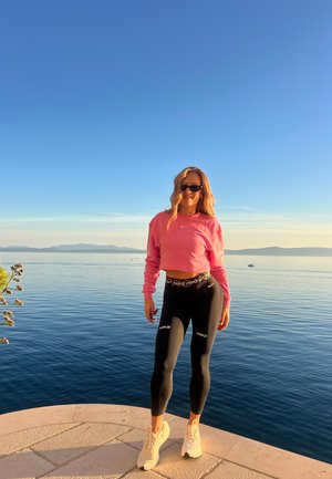 Sudadera corta rosa, leggings Nike Pro negros con acentos blancos, y zapatillas claras, contra un fondo de agua y montañas.