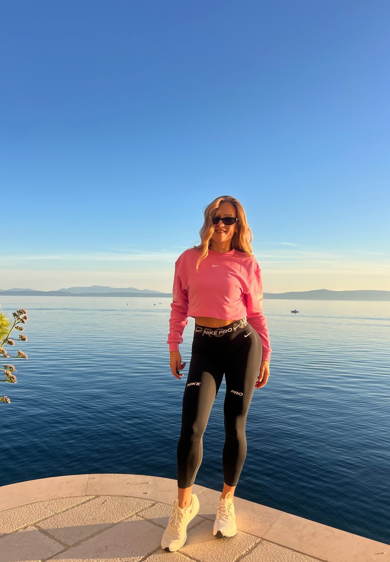 Rosa, verkürzter Sweatshirt, schwarze Nike Pro Leggings mit weißen Akzenten und helle Sneakers, vor einem Hintergrund aus Wasser und Bergen.
