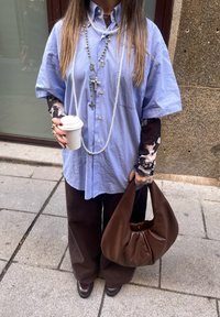 Blue short-sleeved shirt, brown wide-leg trousers, brown leather bag, layered necklaces, coffee cup, patterned arm sleeves.