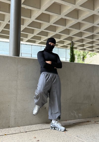 Top negro de manga larga, pantalones cargo de gran tamaño en gris y zapatillas deportivas plateadas con detalles. Persona de pie contra una pared de concreto.