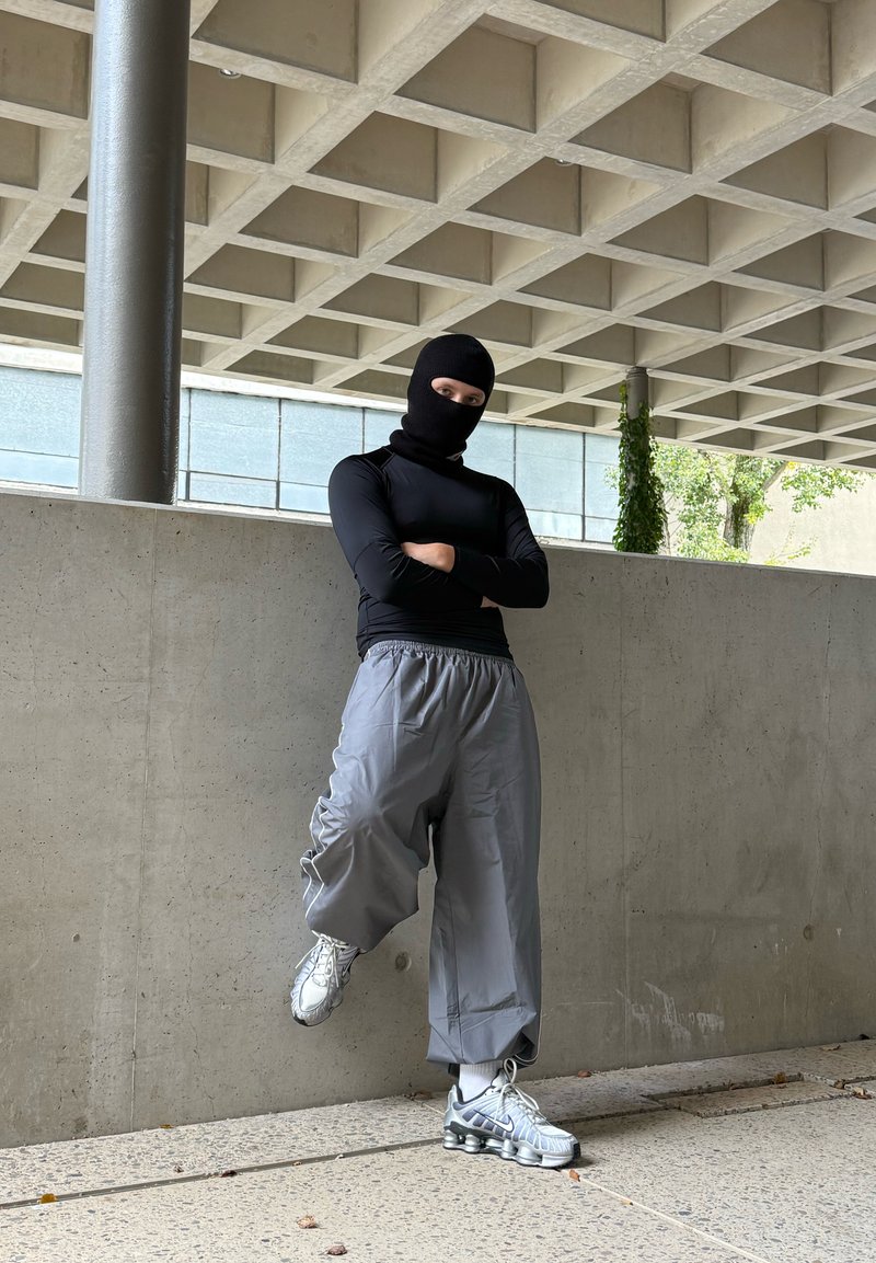 Zwarte lange mouwen top, grijze oversized cargobroek en zilveren sport schoenen met accenten. Persoon staat tegen een betonnen muur.