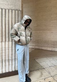 Puhasta jakna u bež boji s uzorkom zelene kamuflaže, s kapuljačom i zatvaranjem na patentni zatvarač, uparena sa sivim trenirkama koje imaju crnu zvijezdu.