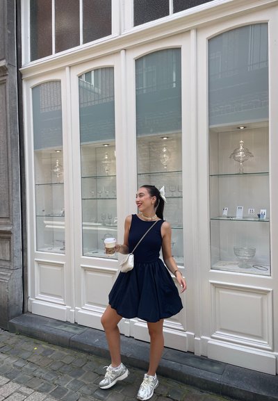 Vestido sin mangas de color azul marino con falda acampanada, zapatillas plateadas, sosteniendo una taza de café. El fondo presenta una vitrina de cristal con artículos de vidrio.