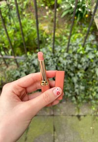 Γυμνό κραγιόν σε χρυσή και ροδακινί θήκη, κρατημένο σε ένα χέρι. Ο πυρήνας διαθέτει λογότυπο και ματ φινίρισμα. Το φό ντο περιλαμβάνει πράσινα στοιχεία.