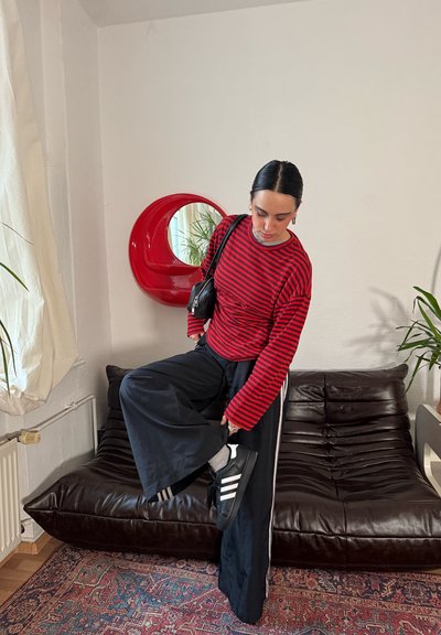Camisa de manga larga a rayas rojas y negras con detalles grises, pantalones negros de pierna ancha y zapatillas negras con rayas blancas, de pie sobre un sofá marrón.