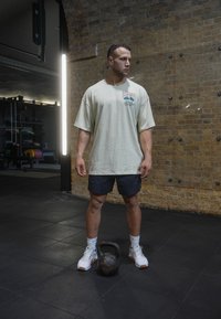 T-shirt oversized bege claro com logotipo, calções pretos, ténis brancos, em pé sobre um piso de ginásio preto ao lado de um kettlebell preto.