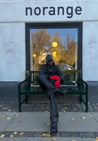 Hombre sentado en un banco verde, vestido con un traje de cuero negro y guantes rojos. Una vitrina muestra luces amarillas redondas y hojas de otoño cercanas.