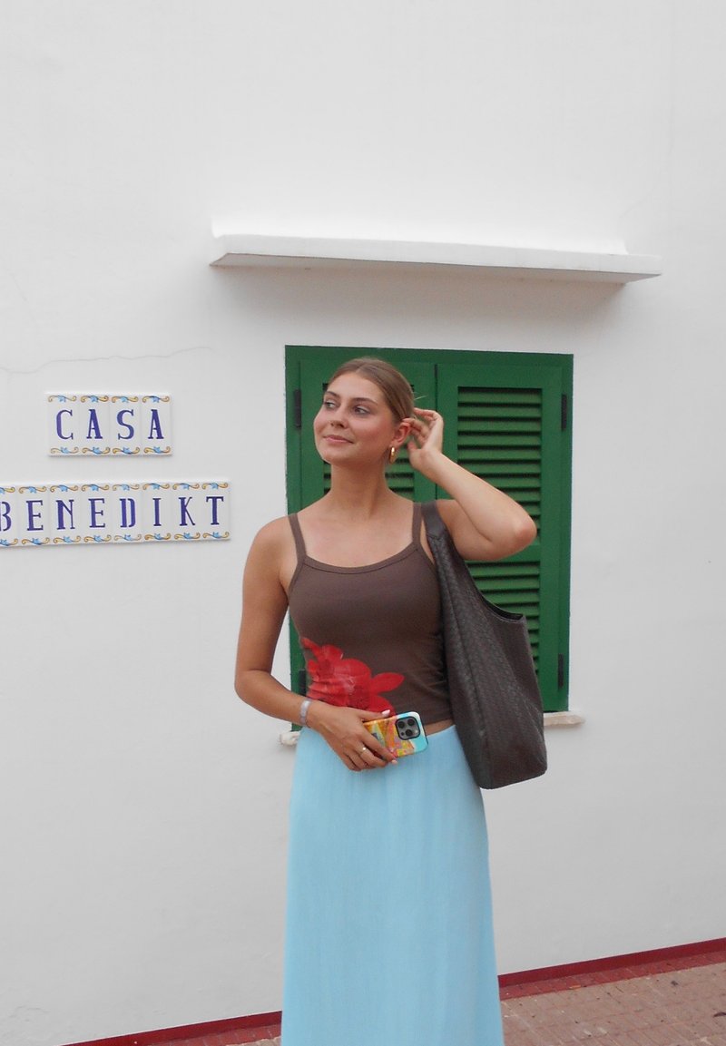 Młoda kobieta w brązowej bluzce i jasnoniebieskiej spódnicy stoi obok białej ściany z oknem z zielonymi okiennicami, trzymając telefon i brązową torbę na ramię.