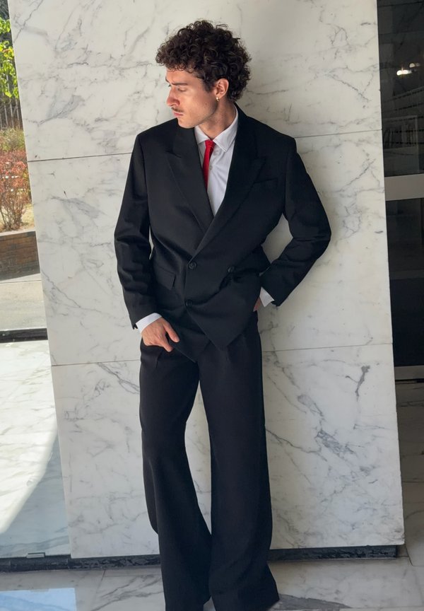 Hombre con cabello rizado que lleva un traje negro de doble botonadura, corbata roja y camisa blanca, de pie contra una pared de mármol blanco mirando hacia un lado.