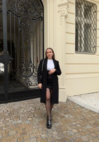 Un cappotto di lana nero sopra un top bianco, abbinato a una minigonna nera e collant trasparenti. Scarpe nere in pelle verniciata e una borsa nera.
