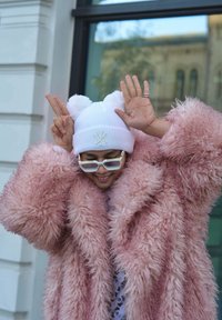 Casaco de pêlo falso rosa fofinho, chapéu de malha branco com pompons, óculos de sol retangulares e transparentes, gestos de mão brincalhões e roupa com padrões por baixo.