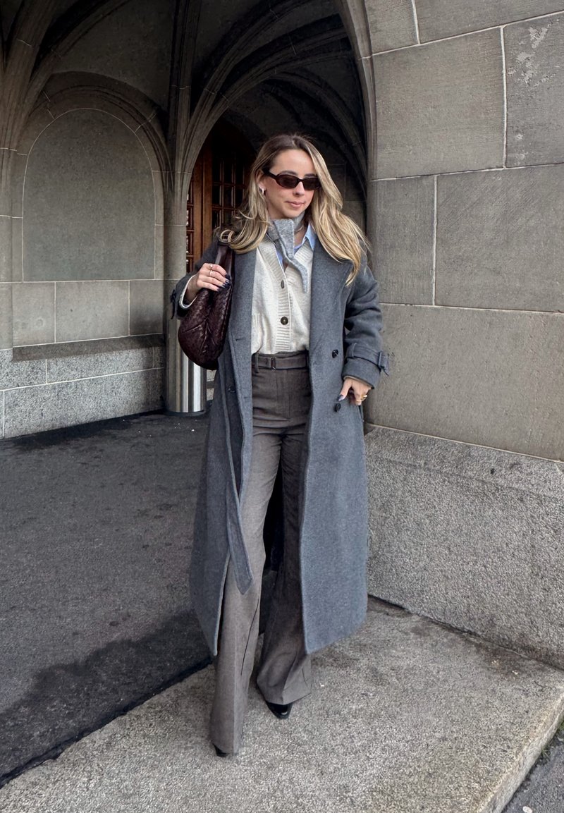 Femme marchant sous une arche en pierre, portant un long manteau gris, un cardigan blanc, un pantalon gris, des lunettes de soleil, et portant un sac à main sombre sur l'épaule.