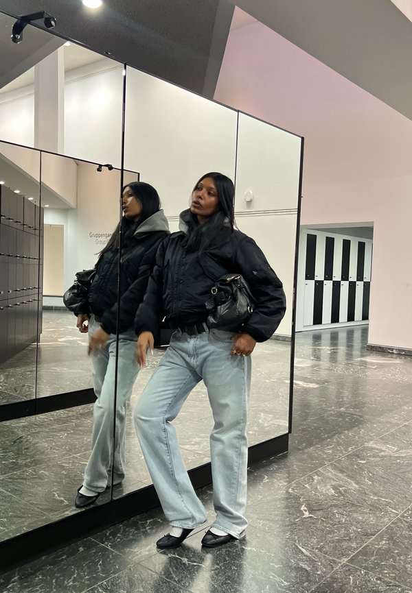 Black puffer jacket, light grey hoodie, light blue jeans, black handbag, and flat shoes. Reflective mirror background in a modern space.