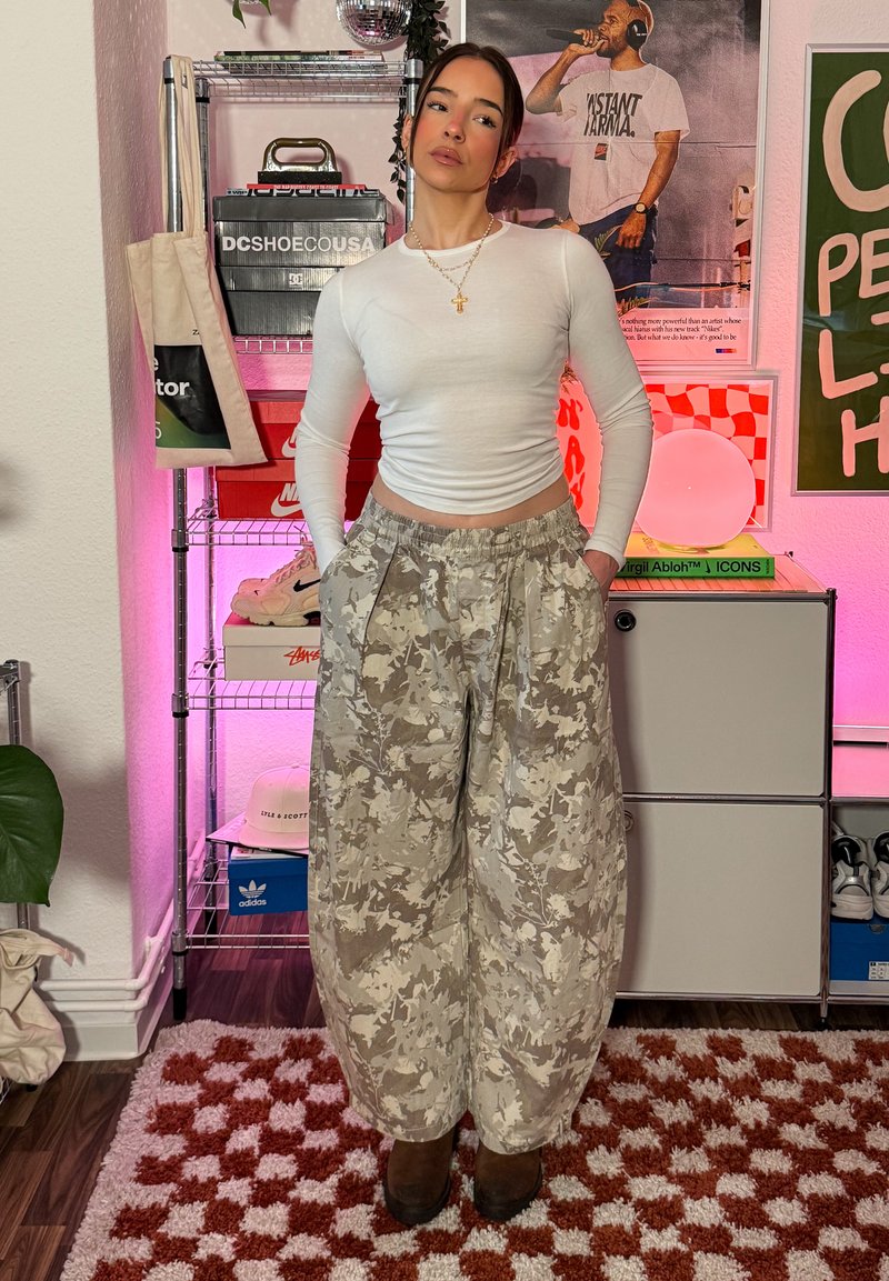 Femme portant un haut blanc à manches longues et un pantalon large beige camouflage, debout les mains dans les poches dans une pièce avec des affiches et des étagères derrière elle.