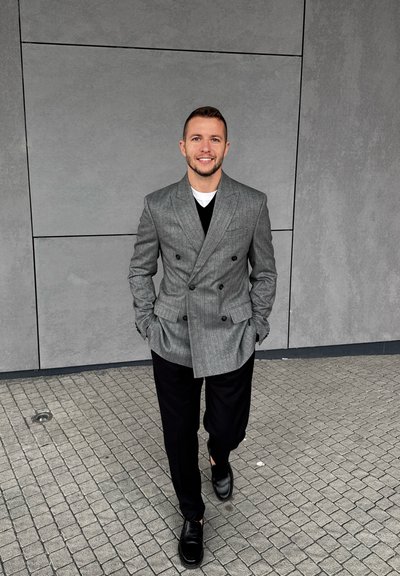 Blazer gris de rayas con doble botonadura sobre una camiseta negra de cuello en V, combinado con pantalones negros y zapatos negros brillantes, caminando sobre una superficie de baldosas.