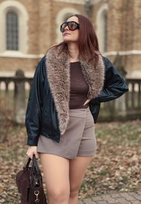 Kožená bomber bunda s kožíškovým límcem, hnědý přiléhavý top, béžové zavinovací šortky a tmavě hnědá kabelka. Modelka má dlouhé červené vlasy a sluneční brýle.