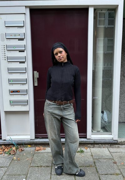 Top negro acanalado con cremallera combinado con pantalones vaqueros grises claros y holgados y zapatos oscuros. De pie contra una pared marrón con buzones y una puerta de cristal.