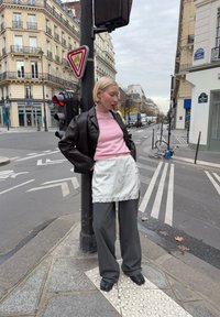 Brown leather cropped jacket, pink jumper, white lace-trimmed apron, grey wide-leg trousers, and black shoes. City street with traffic lights.