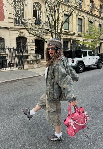 Mujer con chaqueta de camuflaje y pantalones cortos, gorro de piel, gafas de sol, sosteniendo un bolso rosa, pateando con la pierna en una calle de la ciudad con edificios y un coche aparcado detrás.