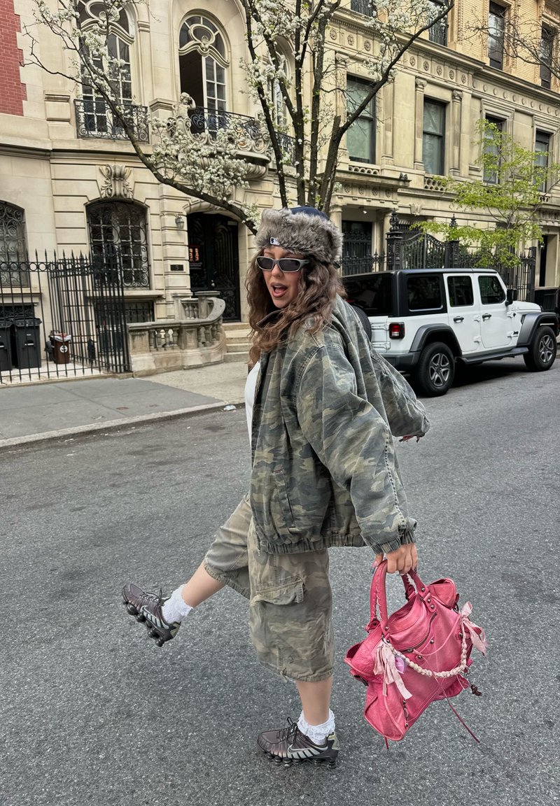 Femme portant une veste camouflage et un short, un chapeau en fourrure, des lunettes de soleil, tenant un sac à main rose, donnant un coup de pied dans la rue de la ville avec des bâtiments et une voiture garée derrière.