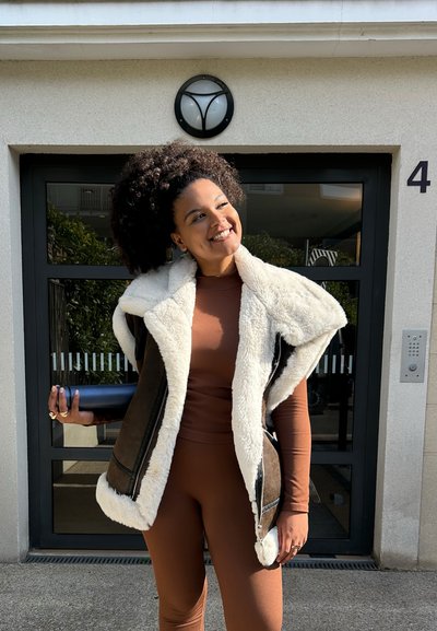 Chaleco de borrego marrón con cuello de piel sintética en color crema, combinado con una blusa marrón de manga larga y leggings ajustados. Sosteniendo un objeto cilíndrico azul.