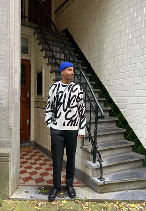 Suéter oversized en blanco y negro con estampado abstracto, combinado con pantalones negros. De pie en unos escalones con un gorro azul y zapatos negros.