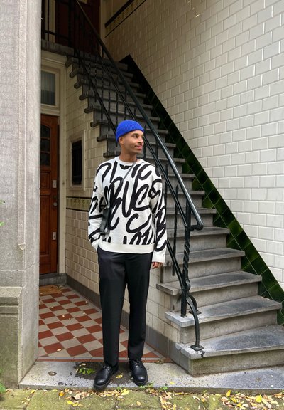 Suéter oversized en blanco y negro con estampado abstracto, combinado con pantalones negros. De pie en unos escalones con un gorro azul y zapatos negros.