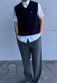 Ναυτικό πλεκτό γιλέκο πάνω από λευκό πουκάμισο με γιακά, σε συνδυασμό με γκρίζες παντελόνες και λευκά αθλητικά παπούτσια. Διακριτό λογότυπο Lacoste στο γιλέκο.