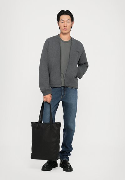 Sudadera gris con cremallera sobre una camiseta verde oscuro, jeans azules y zapatos de cuero negros; sosteniendo una bolsa de tela negra.