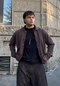 Rudas megztinis su apykakle, po juo juodos spalvos marškiniai, ir ilga pakabuka. Tamsios plisuotos kelnės užbaigia aprangą.