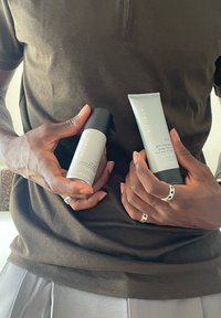 Osoba nosząca pierścionki trzymająca w jednej ręce spray antyperspiracyjny Rituals, a w drugiej balsam do ciała przeciw suchości na tle ciemnej koszuli.