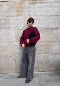 Pull bordeaux, jean gris large, sac noir et chaussures foncées. Un homme se tient contre un mur gris texturé, portant des lunettes de soleil foncées.