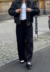 Persona che aggiusta una giacca di denim nera sopra una camicia marrone e un top bianco, indossando pantaloni larghi neri e scarpe bianco e nere su una strada cittadina.