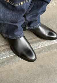Schwarze Leder-Stiefeletten mit spitzer Zehenform, glatter Textur und Blockabsatz, getragen zu dunklen Jeans mit gelben Nähten.
