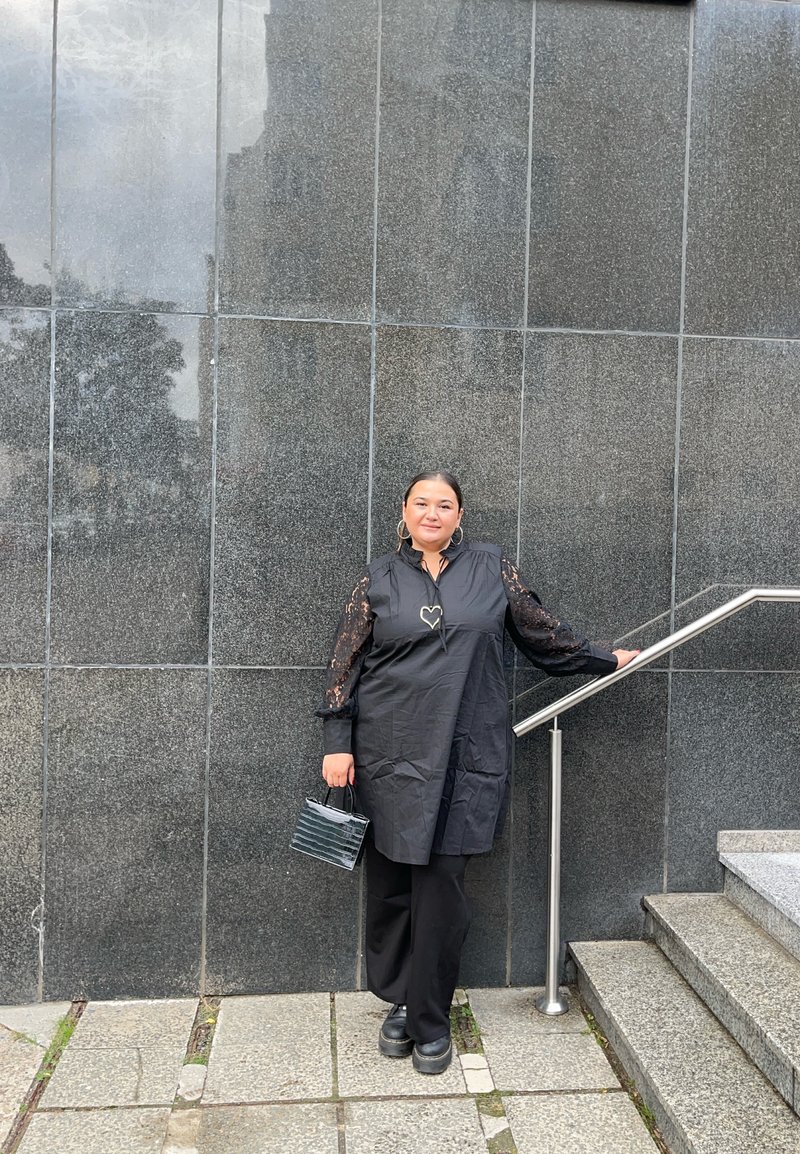 Vestido preto com mangas de renda, um colar em forma de coração e uma bolsa texturizada preta. Apoiando-se em uma parede de pedra e escada. Calças e sapatos pretos.